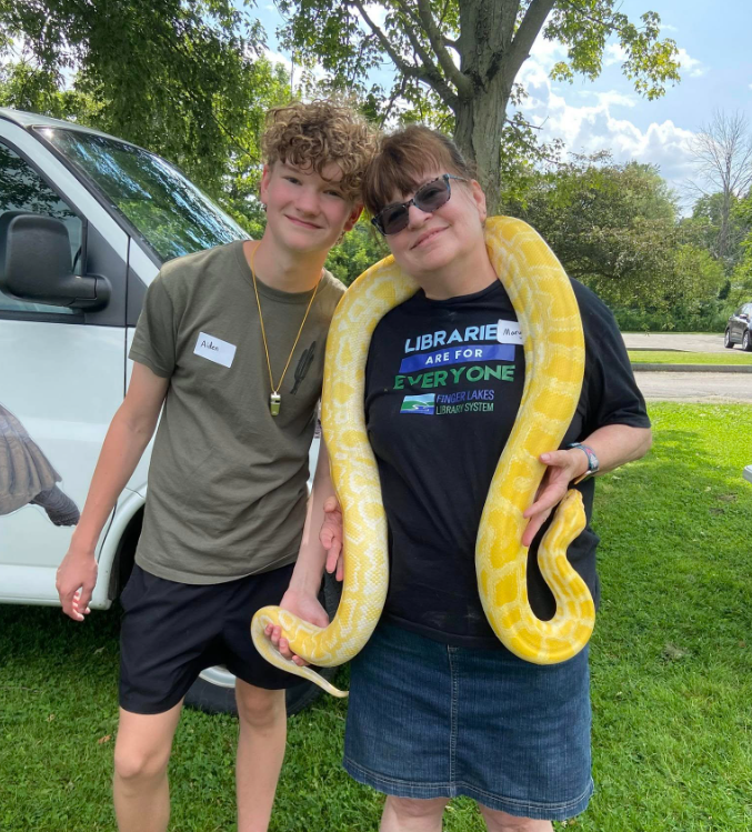a woman holding a large yellow snake smiling next to a teen boy